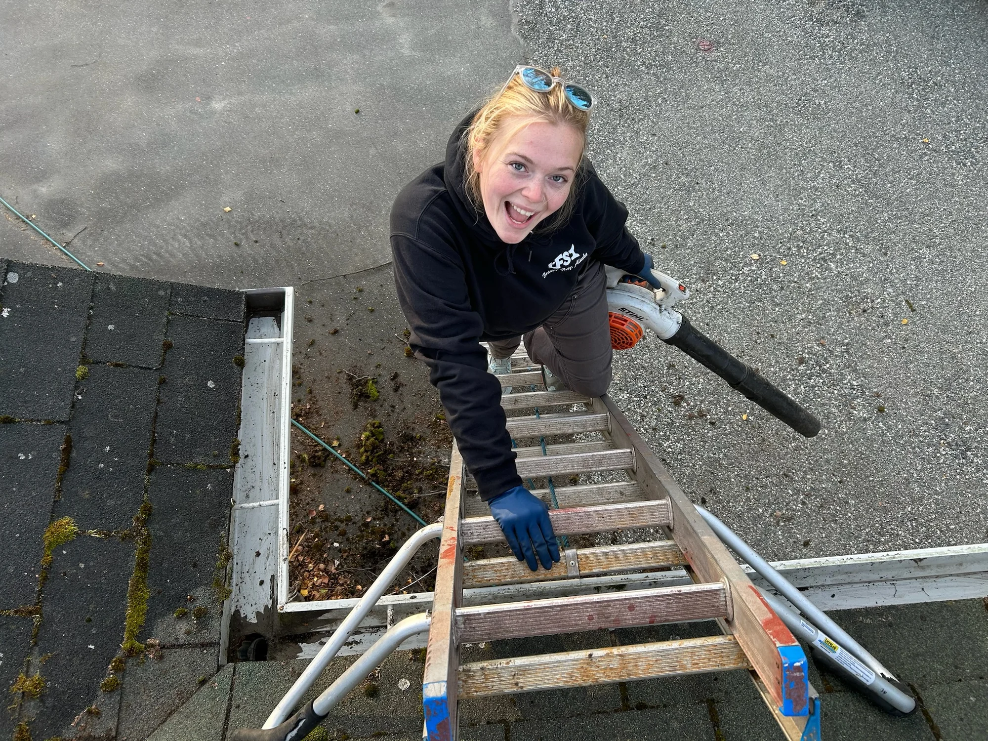 AK Gutter Gals team member smiling on a ladder while clearing gutters